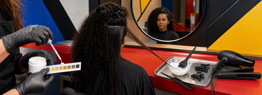 Profissional de salão examinando o rótulo de um produto capilar ao lado de luvas, pincel, tigela e mecha de cabelo